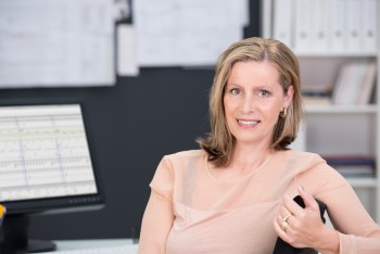 Middle-aged self-employed businesswoman sitting at her desk turning with her arm over the back of her office chair to smile at the camera