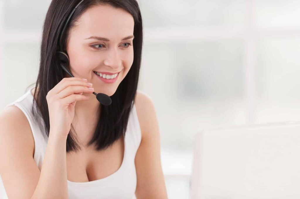 How may I help you? Attractive young woman adjusting her headset and smiling while looking at the laptop