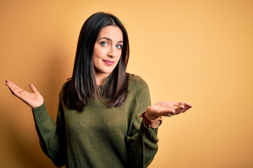 Young brunette woman with blue eyes wearing green casual sweater over yellow background clueless and confused expression with arms and hands raised. Doubt concept.