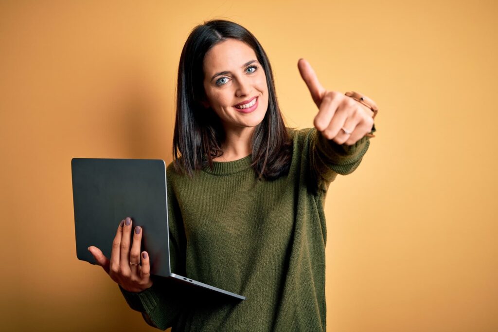 Young brunette woman with blue eyes working using computer laptop over yellow background approving doing positive gesture with hand, thumbs up smiling and happy for success. Winner gesture.